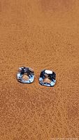 Matched pair of cushion cut natural blue topaz gems displayed on brown textured surface, showing size and clarity details