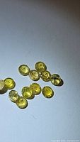Close-up photo of thirteen round cut yellow tourmaline gemstones on a light surface, showing transparency, color and faceting details.