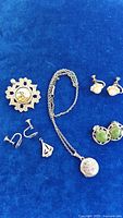 View of sterling silver locket on chain, multiple pairs of sterling earrings including faux pearl, green stone and small silver pairs, and sterling ship charm pendant laid on blue fabric.