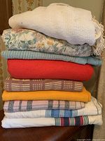Stack of blankets showing different colors and patterns including white, floral, blue, red, plaid, yellow, and striped designs.