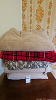 Stack of five blankets showing colors and patterns including beige fleece, red plaid, and digital camouflage.