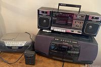 Photo showing three devices: one Sony boombox on top, Sharp 5 CD changer cassette boombox below, and a small Realistic radio on the left side. All on wooden surface.