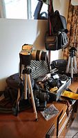 Full lot displaying cameras, tripods, bags and assorted accessories on wooden surface with natural light from window