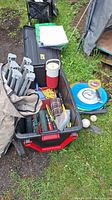 Photo shows an open plastic bin with various camping items including a camp stool frame, red and white cooler, ropes, thermos, toys, and other gear.