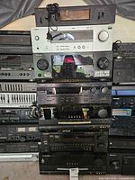 Stack of seven vintage audio electronics stacked vertically including assemblage of amplifiers and receivers with various knobs, displays, and connectors.