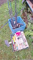 Fishing rods stored upright in a blue crate along with a tackle box filled with lures, lines, and hooks placed on the grass.
