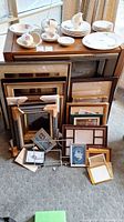 Large assortment of frames stacked and leaning against wood media console; includes some plates and cups on top.