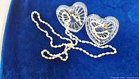Fresh water pearl necklace draped on blue velvet fabric beside a pair of clear Italian crystal heart-shaped trinket bowls.