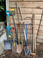 Full view of garden tools including snow shovel, spades, hoe, garden forks, rake, and scraper leaning against wooden shed wall