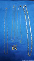 Four gold necklaces and one pair of earrings laid on blue fabric, showing full length and necklace chain styles.