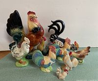 Group photo displaying all 9 ceramic and stone rooster and hen figurines with varied colors and sizes on green surface
