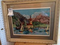 Framed petit point landscape artwork showing boats, church, mountains, and water inside a decorative cream-colored frame.