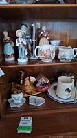 Shelf with various porcelain figurines, decorative mugs, and small collectibles.