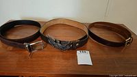 Three men's belts on wooden surface: left black leather belt with silver rectangular buckle, center Harley Davidson brown leather belt with large eagle emblem buckle, right dark brown leather belt with silver buckle.