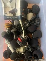 Plastic bin filled with various wired microphones and cables, includes foam windscreens and a packaged USB boom microphone