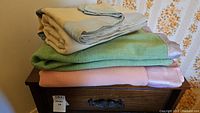 Three folded vintage wool blankets stacked on a wooden furniture piece, colors green, beige, and pink.