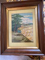 Full view of framed needlepoint depicting a countryside scene of trees, fields, and houses, covered with glass in a wooden frame.