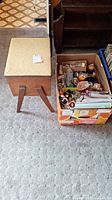Photo showing vintage wooden sewing stool with cushioned beige seat and box of sewing and office supplies placed next to it on carpeted floor.