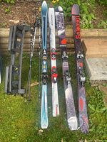 Four pairs of skis with bindings, two sets are Salomon brand, one Head brand pair, with ski poles and a vintage ski rack placed on grass and wooden planks.
