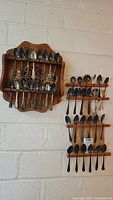 Wide view of three wooden racks mounted on a wall, holding a total of 27 tarnished silver plate souvenir spoons with various ornate handles and bowl designs.