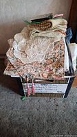 Box containing assorted folded linens including floral patterned fabric, crocheted doilies, and woven placemats.
