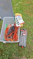 Plastic bin containing orange extension cord, trouble light, sockets, grommets and other small hardware items outside on grass.