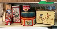 Several vintage tins including a rusted yellow lunch kit with sailing ship image, Edwards Coffee tin, teal and red round tins, and rectangular food tins with child illustrations