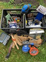 Overall view of the lot showing two plastic crates containing various hand tools, wire brushes, staples, and the Rok handheld sander with sanding discs and leather belt on the grass.