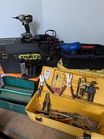 Wide view of Stanley metal toolbox (black with yellow label) with yellow open toolbox holding multiple hand tools including pliers, wire cutters and hex keys. Also visible is a Husky black plastic tool holder on top with other power tools and cords.