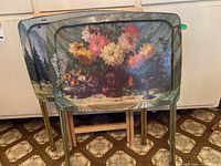 Two vintage metal TV trays with decorative tops showing a still life and landscape scene, leaning against a wall.