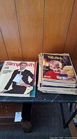 Two stacks of vintage magazines and catalogs on a table against a wood panel wall, showing variety in size and age.