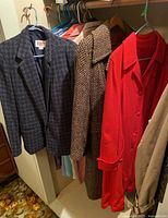 Four vintage coats and jackets hanging on hangers including red long coat, brown herringbone coat, grey blazer, and beige coat