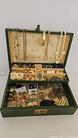 Green jewelry box opened showing hanging necklaces, bangle and top tray compartments of earrings