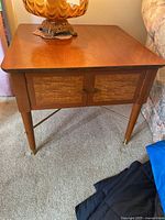 Front view of the vintage MCM solid wood side table showing square top, tapered legs with brass caps, and two cabinet doors with lighter wood grain and brass knobs.