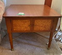 Front view of vintage wooden side table showing front doors with artistic wood grain panels and brass handles, tapered legs with brass caps.