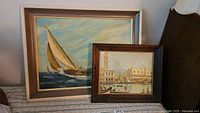 Two framed vintage paintings, one of a sailboat on the water and one of a Venetian canal scene with gondolas, placed side-by-side on a cloth-covered surface.