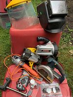 Photo of assorted power tools, welding mask, and flashlight laid on a red surface outdoors, showing Ryobi rotary saw, welding mask with yellow visor, and flashlight.
