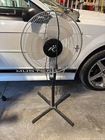 Front view of the 16 inch pedestal fan showing fan blades and protective grille in front of a white car with Mustang logo.