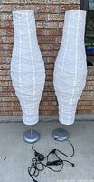 Two floor lamps outdoors against a brick wall. Both have tall white paper shades and silver metal bases with cords and inline switches visible.