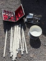 Full lot showing liqueur case with glass bottles, hand crank meat slicer, kitchen scale with bowl and lid, and white metal drawer pulls spread out on gravel surface.