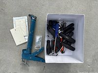 Overview of assorted tools including a blue manual caulking gun, plastic switch plates, small packet of screws, and a box containing multiple hand tools