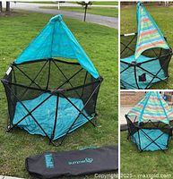 Full view of the hexagonal baby play yard with blue base and striped fabric sunshade, shown outdoors on grass with black Summer carry bag beside it.