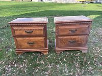 Front view of two wooden bedside tables side-by-side on grass, showing drawer fronts, handles, and trim detail.