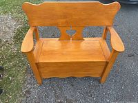 Front view of wooden storage bench showing heart cutout on backrest, storage lid on seat, armrests and scrolled top edge.