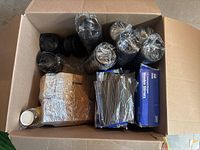 Top-down view into an open box showing several sleeves of black plastic coffee cup lids, two boxes of wooden stir sticks, and two bags of plastic stir sticks, with two cup carriers on the left side.