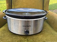 Front view of stainless steel Crockpot slow cooker with black plastic handles and control knob, sitting on green fabric surface.