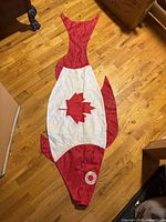 Full view of vintage Canadian fish-shaped flag laid flat on wood floor showing red and white fabric with a large maple leaf in the center.