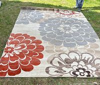 Rug laid flat on grass showing full floral pattern with large blossoms in red, blue-gray, beige, and brown tones on cream background. Edges visible to show dimensions and condition.