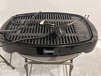 Top-down and angled view of the black Hamilton Beach electric griddle showing the ridged grilling surface, temperature control attached via cord, and power plug.
