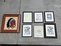 Seven framed needlepoint artworks laid on concrete floor. Large Indigenous chief portrait with feathered headdress in brown and gold-accented frame is distinct. Six smaller framed animal and bird designs in black and brown frames.
